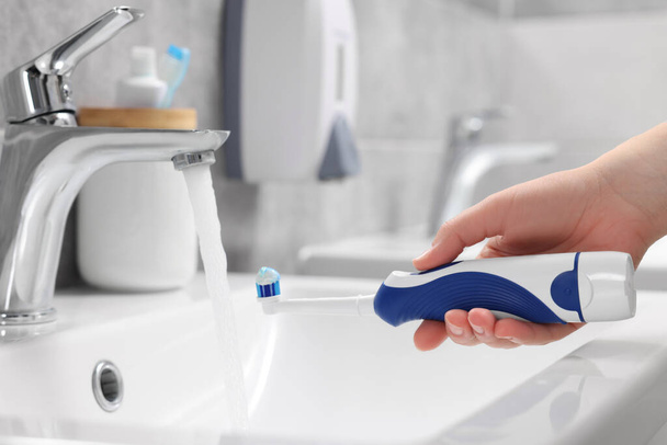 Woman holding electric toothbrush with paste near flowing water above sink in bathroom, closeup - Valokuva, kuva