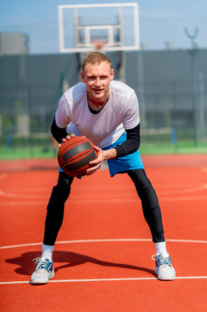Высокий парень баскетболист с мячом показывает свои навыки дриблинга во время тренировки на баскетбольной площадке в парке - Фото, изображение
