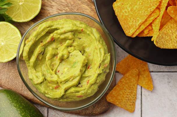 Bowl of delicious guacamole served with nachos chips and lime on white tiled table, flat lay - 写真・画像