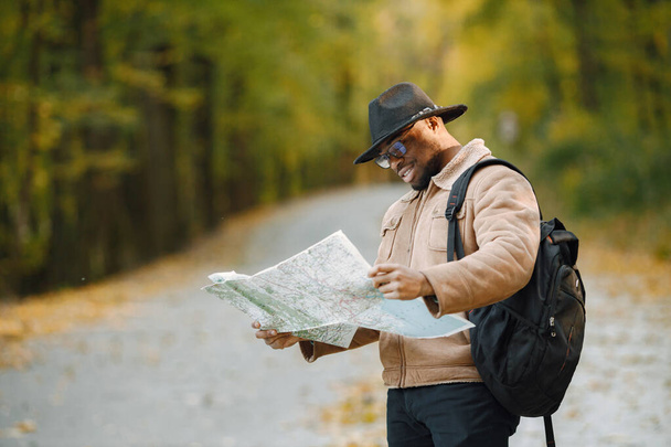 Young black man hitchhiking on road and looking at map. Male traveler feeling lost, traveling alone by autostop. Man wearing brown coat, black hat and backpack. - Photo, image