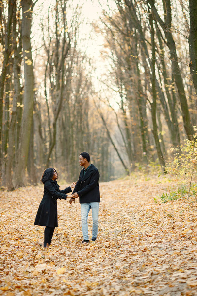 Black young man and his girlfriend holding hands. Romantic couple walking in autumn park. Man and woman wearing black coats. - Fotó, kép