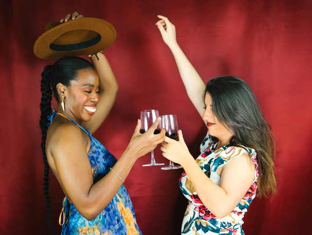 Una ragazza africana e italiana che balla e beve vino felice giocando insieme con un cappello marrone - Foto, immagini