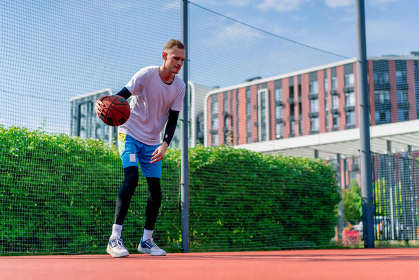 Ψηλός μπασκετμπολίστας με την μπάλα δείχνει τις ικανότητές του ντρίμπλα κατά τη διάρκεια της προπόνησης στο γήπεδο μπάσκετ στο πάρκο - Φωτογραφία, εικόνα