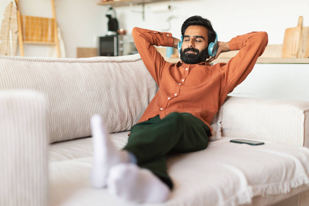 Zenei kikapcsolódás szabadidő. Közel-keleti férfi pihen a fülhallgató élvezi kedvenc zene és dalok fekvő kanapén a kezét a fej mögött, pózol csukott szemmel otthon - Fotó, kép
