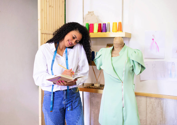 Kıyafet işi. Başarılı Giyim Tasarımcısı Cep telefonuyla iletişim kuruyor ve not alıyor, Couture Atıcısının iç mekanında müşteriler için kıyafet yaratıyor, Kukla ve Sohbetin yanında duruyor - Fotoğraf, Görsel