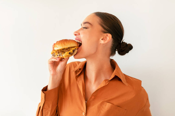 Mujer joven satisfecha mordiendo sabrosa hamburguesa con placer, disfrutando de la comida engañosa sobre fondo blanco de la pared. Señora pidiendo almuerzo para comida para llevar entrega en restaurante de comida rápida - Foto, imagen