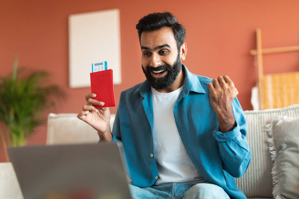 Viajante árabe Guy com passaporte e bilhetes olhando para laptop e gesto sim, cheio de alegria e emoção, comemorando férias e websurf on-line grande oferta agência de viagens em casa - Foto, Imagem