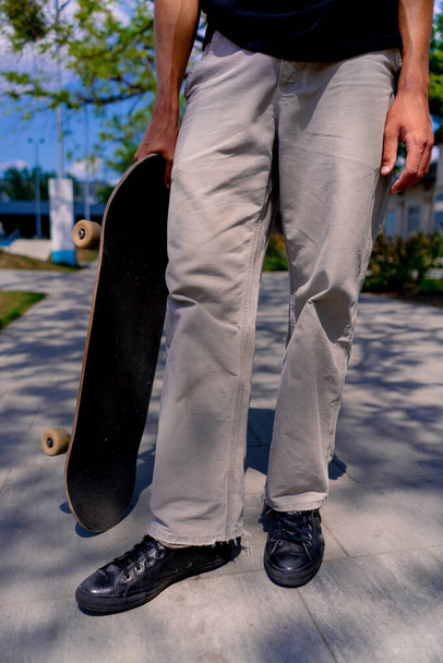 Gros plan sur les jambes du skateboarder en jeans et un skateboard tenu par un jeune homme dans le skatepark de la ville - Photo, image
