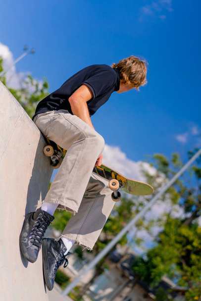 A young guy skateboarder with long hair sits on his skateboard on the edge of a skate pool at city skatepark - Foto, Bild