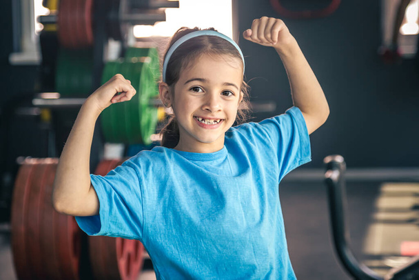Очаровательная маленькая девочка показывает свои бицепсы и улыбается во время тренировки в спортзале. - Фото, изображение