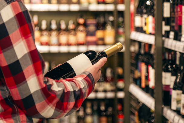 Mężczyzna trzymający butelkę wina w monopolowym sklepie z winem, wybierający właściwe wino ze wszystkich odmian butelek wina na półkach. - Zdjęcie, obraz