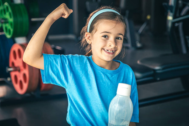 Charming little girl is shows her biceps and smiling while working out at gym. - Photo, Image