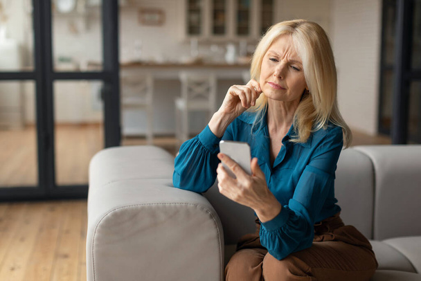 Bad news concept. Confused mature woman holding smartphone, looking at cellphone screen with worried expression, reading unplesant message, sitting on couch, copy space - Photo, Image