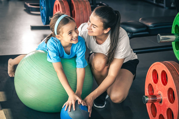 Klein sportief meisje en jong mooi moeder doen stretching met fitness bal in de sportschool. - Foto, afbeelding