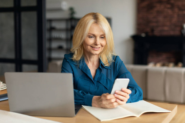 Home business success. Happy mature female entrepreneur with laptop and phone sitting at desk in living room. Cheerful businesswoman working from home - Foto, afbeelding