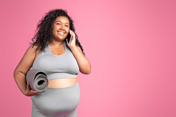 Χαμογελώντας μαύρο συν μέγεθος γυναίκα ανάπαυσης μετά την προπόνηση, κρατώντας fitness mat και μιλώντας στο κινητό τηλέφωνο, κοιτάζοντας χώρο αντίγραφο πάνω από ροζ φόντο στούντιο - Φωτογραφία, εικόνα