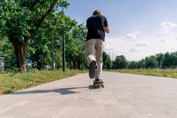 Junger Skater fährt Skateboard auf dem Weg durch den Stadtpark vor dem Hintergrund von Bäumen und Himmel - Foto, Bild