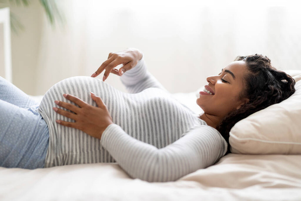 Retrato de mujer embarazada negra caminando con los dedos acroos su gran vientre mientras está acostada en la cama en casa, mujer embarazada sonriente preparándose para la maternidad, disfrutando de su tiempo de embarazo, vista lateral - Foto, imagen
