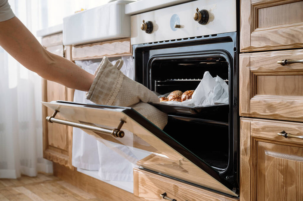 обрезанный снимок женской руки в перчатке достает поднос для выпечки с домашним хлебом из электрической духовки на домашней кухне. Концепция приготовления - Фото, изображение