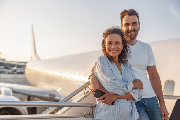 Porträt eines glücklichen Touristenpaares, Mann und Frau, die aufgeregt aussehen, während sie zusammen im Freien stehen und bereit sind, bei Sonnenuntergang das Flugzeug zu besteigen. Urlaub, Lifestyle, Reisekonzept - Foto, Bild