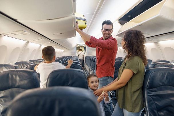 陽気な男は笑顔で妻を見て、飛行機で家族と一緒に旅行中に荷物をコンパートメントに運ぶ。家族の休暇、交通概念 - 写真・画像
