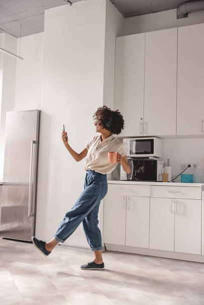 Full-length φωτογραφία του ευτυχισμένη όμορφη γυναίκα κρατώντας κύπελλο, ενώ χορεύουν στην κουζίνα και χρησιμοποιώντας ακουστικά - Φωτογραφία, εικόνα
