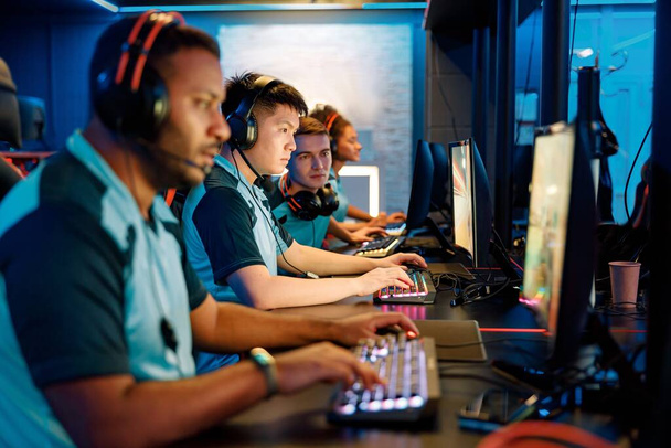 Grupa multietnicznych graczy cybersportu w słuchawkach siedzi przed komputerami i gra w gry wideo online w klubie komputerowym - Zdjęcie, obraz