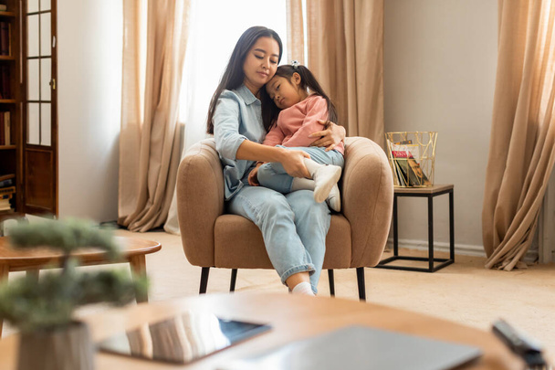 Snuggle Time. Asian mother holding little daughter in arms sitting in armchair, cuddling and snuggling up together for warmth and affection at home interior. Motherhood Bonding Moments Concept - Foto, Imagem
