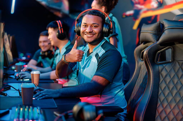 Happy young man expressing satisfaction with game while giving thumbs up gesture during participation in international gaming event in gaming club - Photo, Image