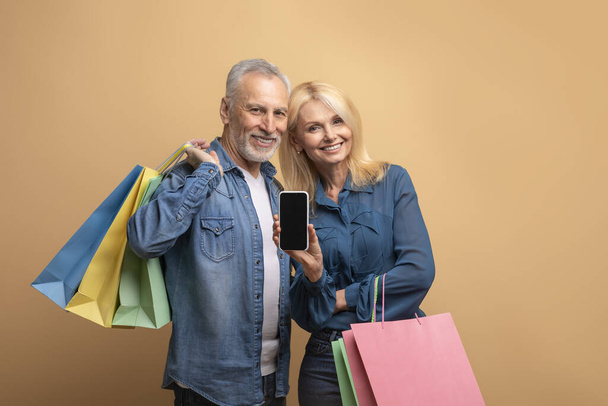 E-commerce, online sale. Happy cheerful beautiful senior couple grey-haired man and blonde woman with colorful shopping bags and smartphone in their hands isolated on beige background, copy space - Фото, зображення
