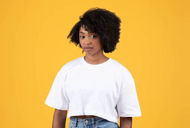 Unhappy serious pretty young black curly lady in white t-shirt look at camera, isolated on orange background, studio. Contempt, disgust, bad news reaction and human emotions - Valokuva, kuva