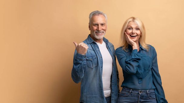 Excited cheerful beauitufl elderly man and woman in casual denim outfit isolated on beige background, pointing at copy space. Happily married senior couple showing great offer, advertisement - Foto, Imagem