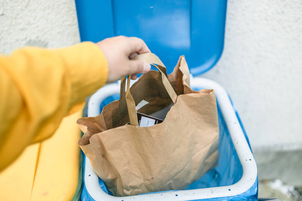 Blanke vrouw sorteren van afval, het gooien van een gebruikte papieren zak in een kleine prullenbak in de buitenlucht in de buurt van een huis  - Foto, afbeelding