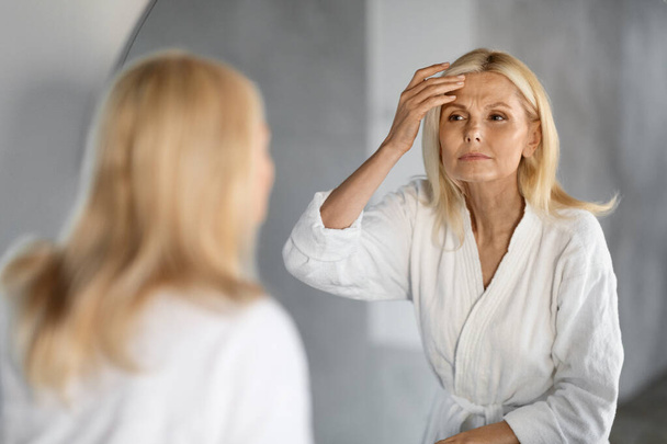 Ώριμη γυναίκα κοιτάζοντας στον καθρέφτη και αγγίζοντας ρυτίδες στο πρόσωπό της, αναστατώσει όμορφη ηλικιωμένη γυναίκα Εξετάζοντας λεπτές γραμμές στο μέτωπο, υποφέρουν γήρανση του δέρματος, επιλεκτική εστίαση στην αντανάκλαση, Closeup - Φωτογραφία, εικόνα