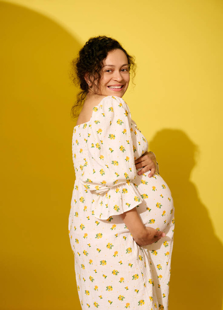Göndör, többnemzetiségű terhes nő babát vár, széles mosollyal néz a kamerába, elszigetelve a sárga stúdió hátterétől. Fogamzóképes. Szülés. Gyönyörű terhesség és szülési szabadság fogalma - Fotó, kép