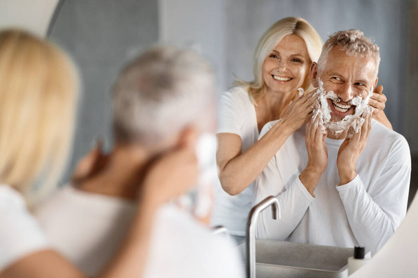 Šťastný starší pár baví při přípravě spolu v koupelně, veselý starší manželé stojící v blízkosti zrcadla, žena objímající manžela při holení, selektivní zaměření na reflexi - Fotografie, Obrázek