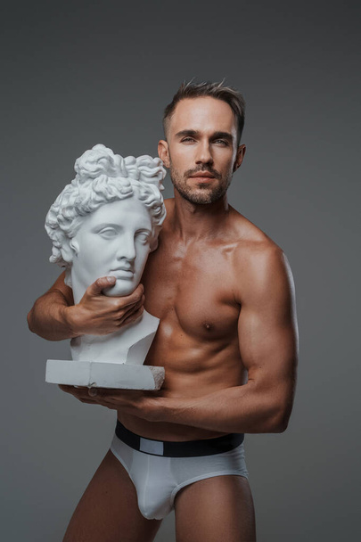 Очаровательный мужчина-модель, одетый только в нижнее белье, обнимает бюст греческой скульптуры, источая очарование и очарование, с серым фоном - Фото, изображение