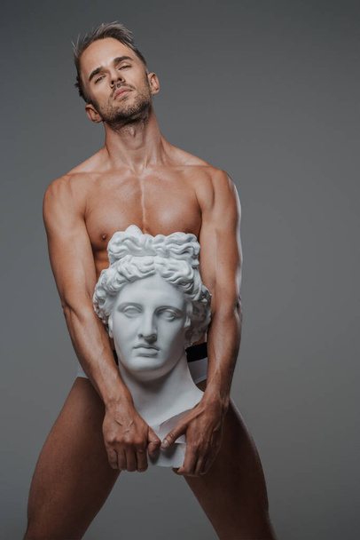 Харизматичный мужчина-модель со скульптурным телосложением позирует рядом с бюстом древнегреческой скульптуры - Фото, изображение