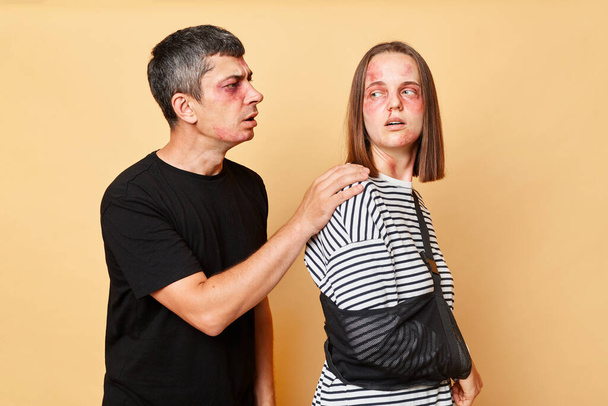 Brunette man and woman with bruise isolated over beige background husband asking for forgiveness from his wife after being severely beaten during family conflict - Foto, Imagen