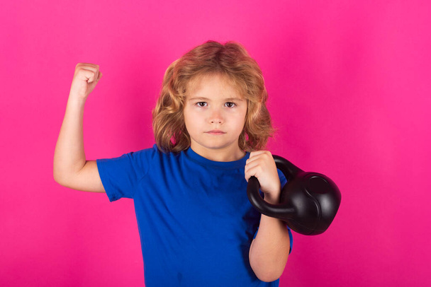 Kid with kettlebell in studio on red pink background. Sport activities at leisure with children. Blonde boy holding dumbbells. Sports exercises for children. Funny child lifting the kettlebell - Photo, Image