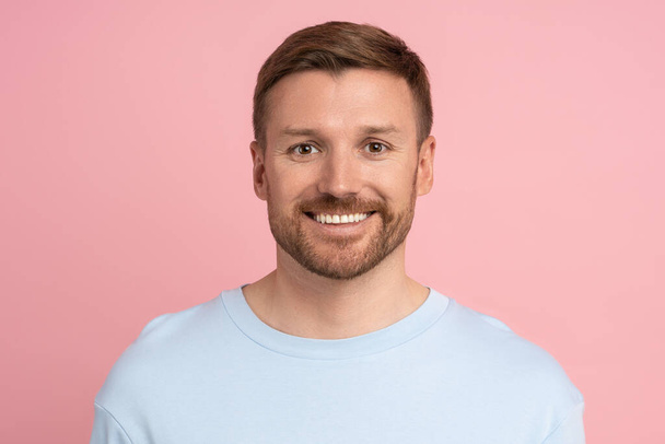 Amerikai szakállas fiatalember portréja, amint a kamerát nézi, amint rózsaszín háttérrel mosolyog. Pozitív középkorú férfi érzés öröm, optimizmus. Vidám, nyugodt fickó, élvezi az életet. Őszinte emberi érzelmek. - Fotó, kép