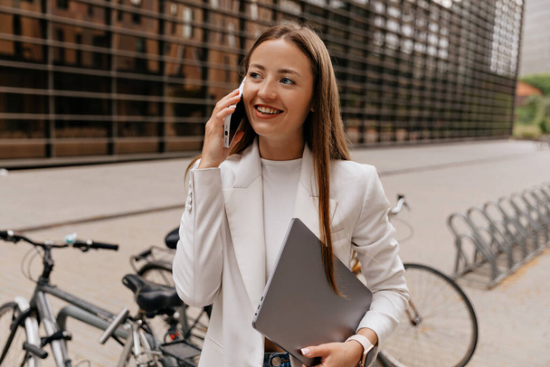 Houkutteleva tyylikäs eurooppalainen nainen valkoisessa takissa ja paidassa puhuu puhelimessa, pitäen kannettavaa tietokonetta yrityskeskusta vastaan. Käsitteen elämäntapa, teknologia nyky-yhteiskunnassa.  - Valokuva, kuva