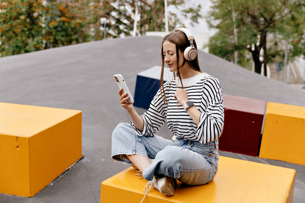 Foto completa de menina bonita elegante com cabelo solto vestindo camisa listrada e jeans está usando smartphone e ouvir música ao ar livre no dia de verão. - Foto, Imagem