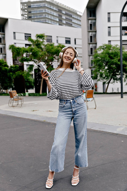 Полнометражное фото стильной красивой девушки в джинсах и летней рубашке на каблуках держит смартфон и наслаждается музыкой в наушниках на улице в городе - Фото, изображение