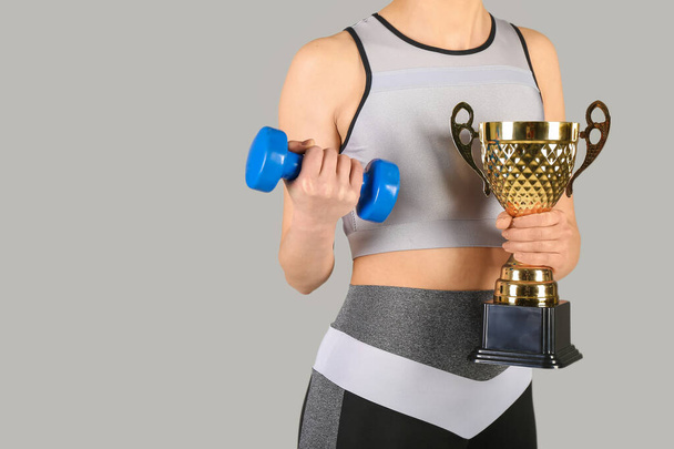 Sporty young woman with dumbbell and gold cup on grey background, closeup - Photo, Image