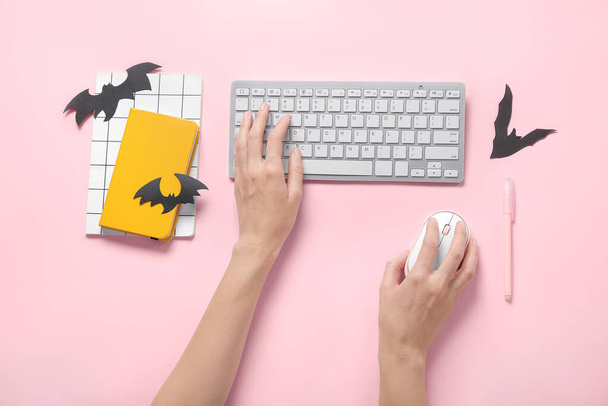 Female hands with modern computer keyboard, mouse, notebooks and paper bats for Halloween on pink background - Photo, Image