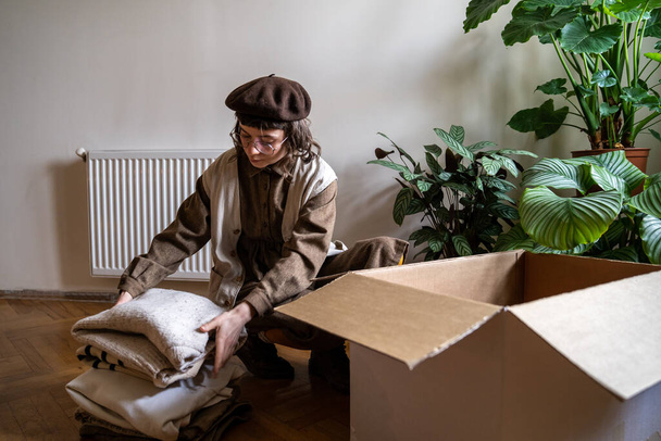 Eco activist hipster girl stacking old clothes into cardboard box at home, sorting unwanted items to recycle. Clothing waste and recycling, conscious living concept. Donation, second hand, save planet - Photo, Image