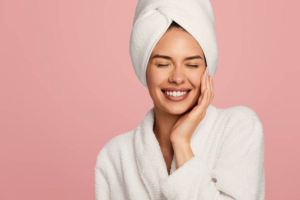 Joyeux jeune femme en peignoir et serviette sur la tête toucher la peau du visage après la procédure de bain debout sur fond rose - Photo, image