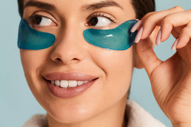 Crop young female applying blue color rejuvenating eye patches with toothy smile - Photo, Image