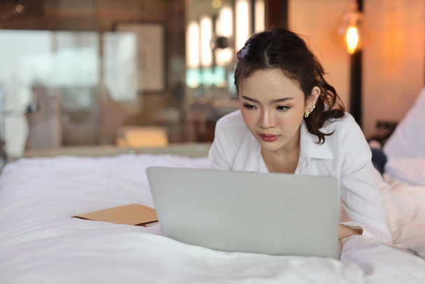Aktivní asijská žena ležící na posteli a pracující na počítači s notebookem v hotelu, krása podnikatelka s ložnicí pozadí. Koncept technologie životního stylu - Fotografie, Obrázek